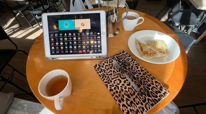 A tablet displaying app icons sits on a wooden café table alongside a cup of tea, a latte, and a pastry on a leopard-print placemat, capturing a casual work session that pairs well with the post about attending a Rialo BuilderHub discussion on "Latch," a developer access management tool for AI providers.