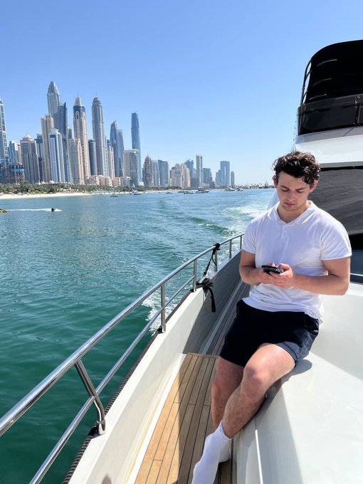 A young man in a white polo shirt and black shorts sits on a yacht using his phone, with the Dubai Marina skyline in the background.