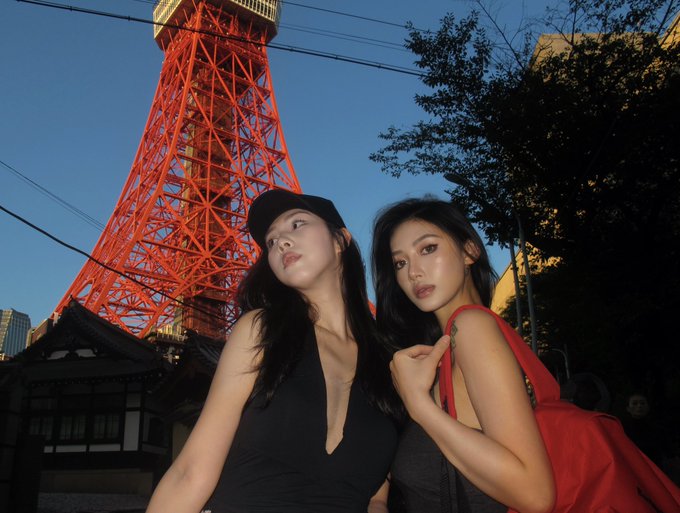 Two stylish women pose confidently in front of Tokyo Tower's iconic red structure, with one wearing a black cap and top, and the other carrying a vibrant red bag. The image serves as a polished lifestyle visual accompanying a tweet correcting misconceptions about Backpack, framing the exchange to its audience with an air of sophistication and international appeal.