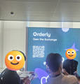 A photo taken at an event showing a banner for 'Orderly Network' and 'Merlin Chain' with QR codes and a person in the foreground.