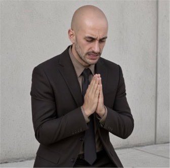 A bald man in a dark suit with his hands pressed together in a prayer position, eyes closed, standing against a plain gray wall.