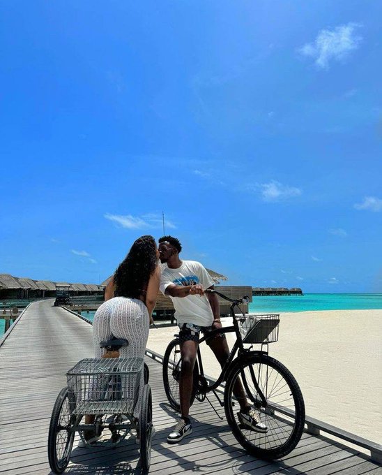 A couple kissing on a wooden boardwalk over turquoise water at a tropical resort, one person on a bicycle and the other in a wheelchair, under a bright blue sky.