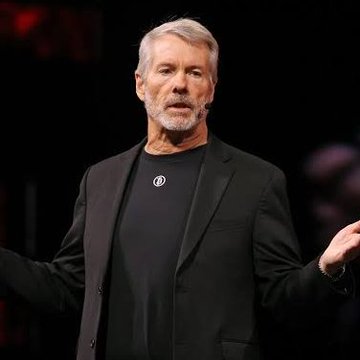 An older man with gray hair and beard wearing a black jacket stands on a stage with his arms slightly outstretched, speaking to an audience.