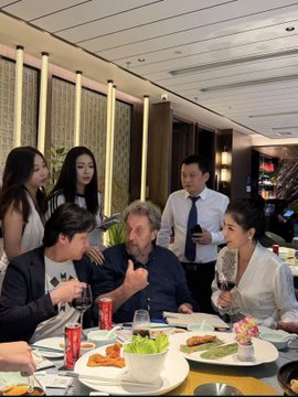 A group of people, including an older man with a beard, are gathered around a restaurant table sharing a meal, with a Coca-Cola can visible among the dishes.