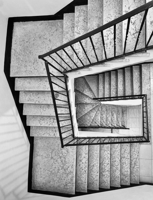 A black and white photograph of a spiral staircase with marble steps and a metal railing, viewed from above.