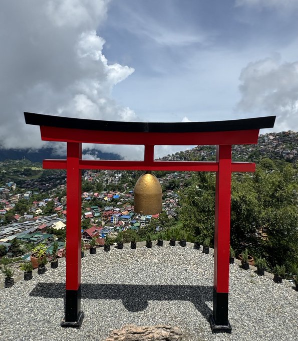A large red torii gate frames a hillside cityscape and a golden egg-shaped structure beneath a cloudy sky.
