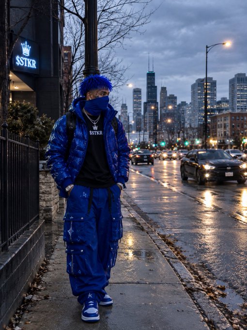 A young man with blue hair wearing a matching blue puffer jacket, pants, and face covering stands on a city street with Chicago skyline visible in the background near an "SSTKR" storefront.