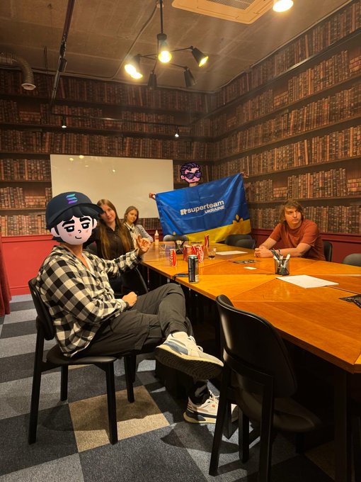 A group of people gathers around a conference table in a library-like room with bookshelf wallpaper, where one person holds up a Superteam Ukraine flag and another has their face obscured with a cartoon character emoji overlay, capturing the friendly atmosphere of a Solana community meetup.