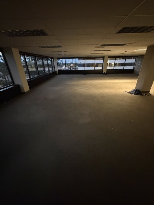 A dimly lit, empty office space with large windows showing an outdoor view, carpeted floors, and a paper towel dispenser near a column.