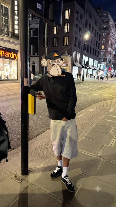 A person wearing a monkey face mask, a black long-sleeve shirt, gray shorts, and sneakers leans against a pedestrian crossing pole on a city street at night.
