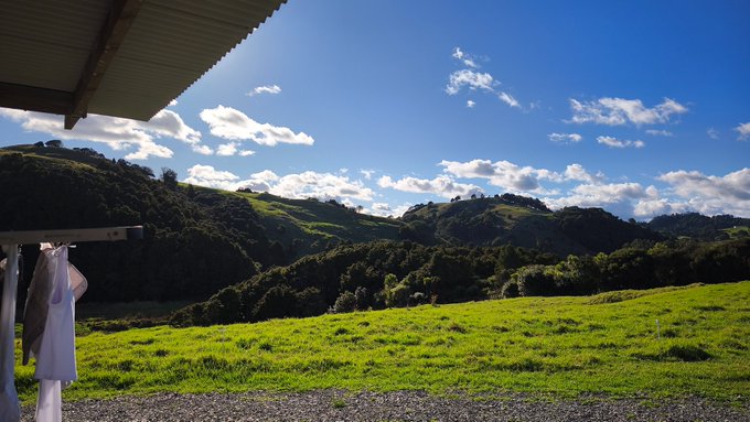 A scenic landscape view from under a roof shows rolling green hills and a bright blue sky with scattered clouds.