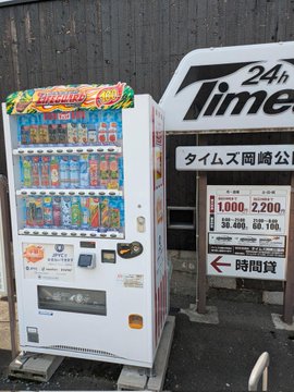 A white "JPYC" vending machine stocked with colorful beverages stands beside a "Times 24h" car rental sign displaying parking rates of "1,000/2,200" and a 24-hour parking notice, marking this as the second location found that accepts JPYC payments.