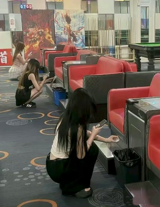Several young women crouch on the patterned carpet of what appears to be a cinema lobby, looking at their phones near red and black booth seating, with a digital clock reading "07:00" visible in the background.