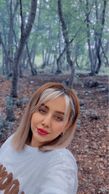 A woman with a blonde and brown bob haircut and red lipstick takes a selfie in a forest with bare trees and brown leaves on the ground.
