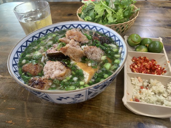 A bowl of Vietnamese noodle soup, likely bún chả or a similar dish, served with fresh herbs, lime, chili, and garlic on a wooden table.