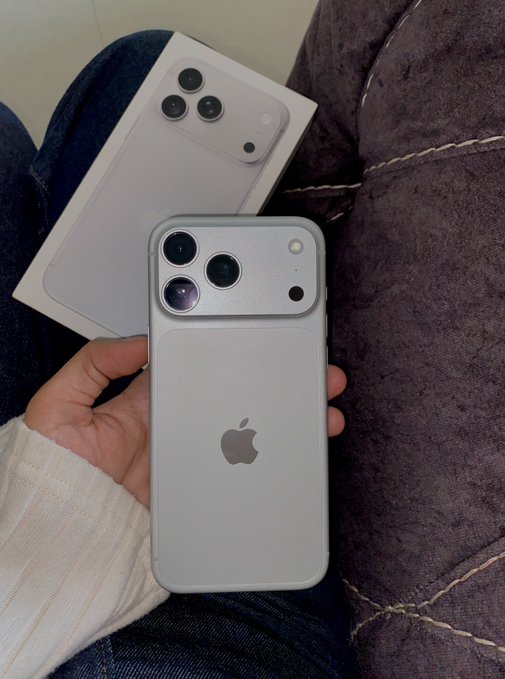 A hand holds a silver/blue iPhone Pro model alongside its white Apple retail box, resting on a denim-covered lap next to a dark brown couch.

The image relates to "thank you bitcoin" by celebrating a recent purchase, suggesting the cost of the premium smartphone was funded by cryptocurrency earnings.