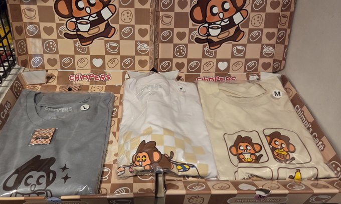 An open cardboard box labeled "CHIMPERS" reveals three folded t-shirts: a grey one with "L" tag, a white one with a checkered design, and a cream one with "M" tag featuring a cartoon monkey.