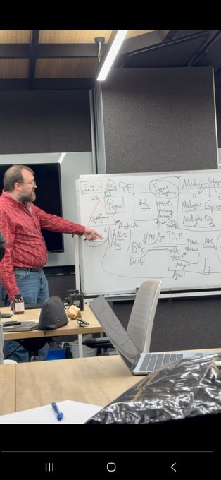 A man in a red plaid shirt stands in front of a whiteboard covered in technical diagrams, explaining concepts that include references to "VPN," "DeFi," and "Multiple Engines," which aligns with @MidnightNtwrk's announcement of being the "VPN for DeFi."