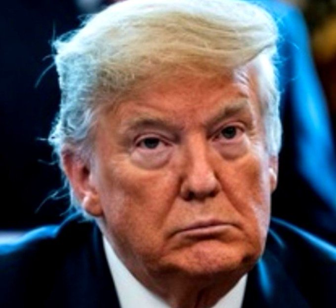 A close-up portrait of Donald Trump wearing a dark suit with a serious expression against a dark blue background.