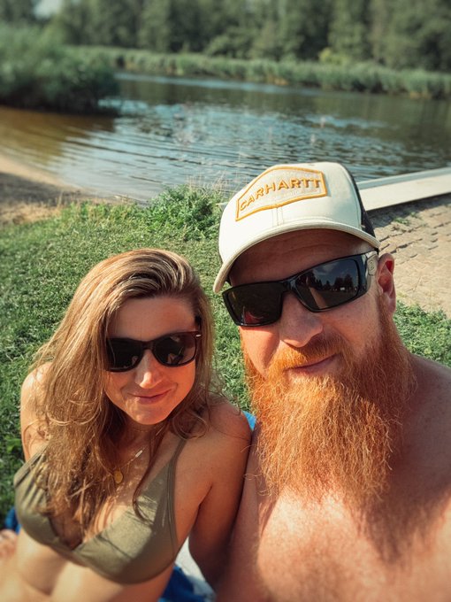 A selfie of a couple by a riverbank; the man has a large red beard, wears an "EARHART" trucker cap and sunglasses, while the woman beside him wears sunglasses and an olive bikini top.
