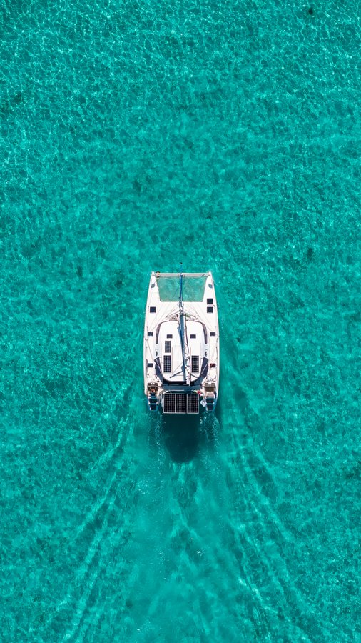 An aerial view of a white catamaran anchored in crystal-clear turquoise waters, likely representing the 56th mint in a limited collection of 69, as referenced in the post. The boat's wake in the pristine water symbolizes the growing rarity as the collection approaches its full size.