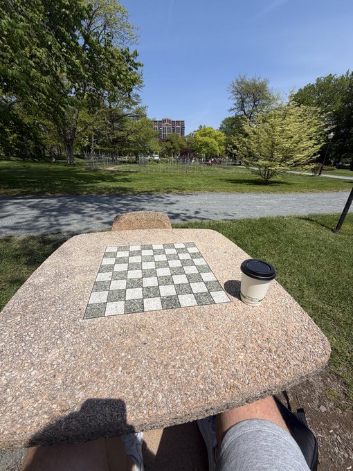 A man's legs are visible underneath a table with a chess board, and there is a cup of coffee next to the board. There is also a person standing behind the table who appears to be watching the game. The post suggests that someone wants to play checkers or chess but has not yet started.