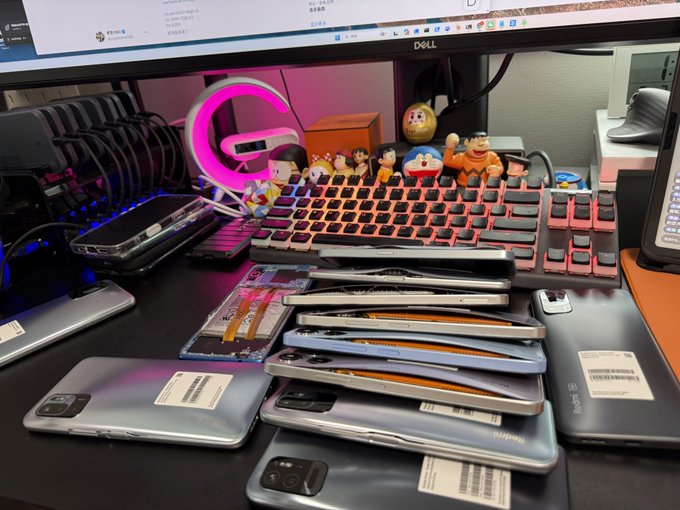 A messy desk displays a stack of used smartphones with visible battery swelling, alongside a Dell monitor, RGB keyboard, and small figurines, with a note mentioning "redmi" visible on one device. The image illustrates the user's frustration about phone batteries bulging, referencing @grass and demanding refunds for the defective devices.