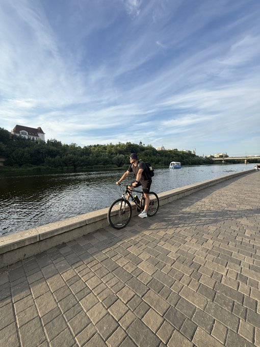 A man wearing a cap, dark t-shirt, and backpack stands with a bicycle on a paved riverside walkway under a partly cloudy blue sky, with trees and a bridge visible in the distance.