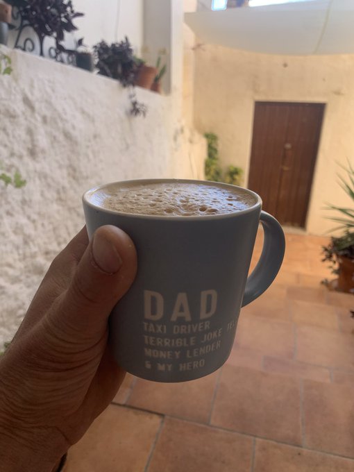 A hand holds a gray coffee mug labeled 'DAD - TAXI DRIVER, TERRIBLE JOKE TELLER, MONEY LENDER & MY HERO' filled with a frothy beverage, set against an outdoor courtyard.