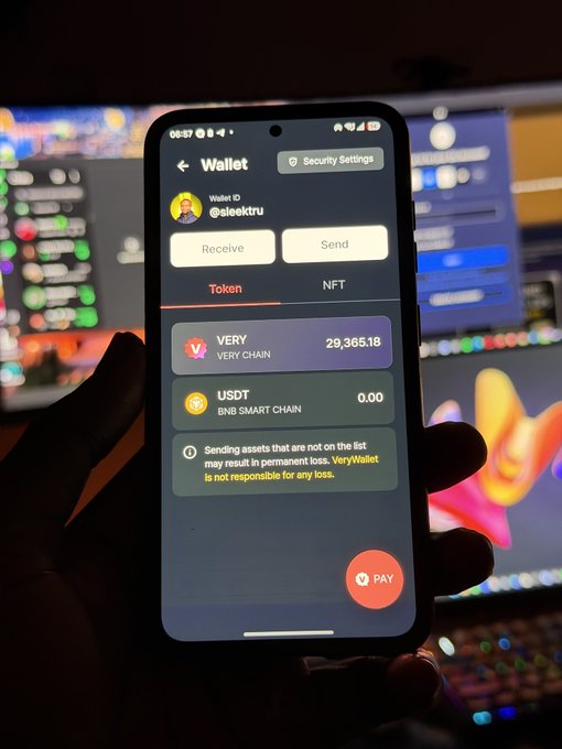 A hand holds a smartphone displaying a crypto wallet interface for '@sleektru' showing a balance of 29,365.18 VERY tokens on the VERY CHAIN, with trading screens visible on a computer monitor in the background.