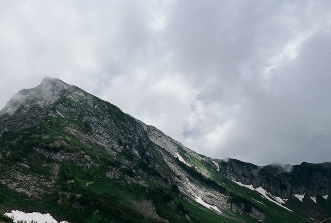 A dramatic mountain landscape featuring rocky peaks partially shrouded in low-hanging clouds and mist, with green grassy slopes and patches of snow under an overcast sky.
