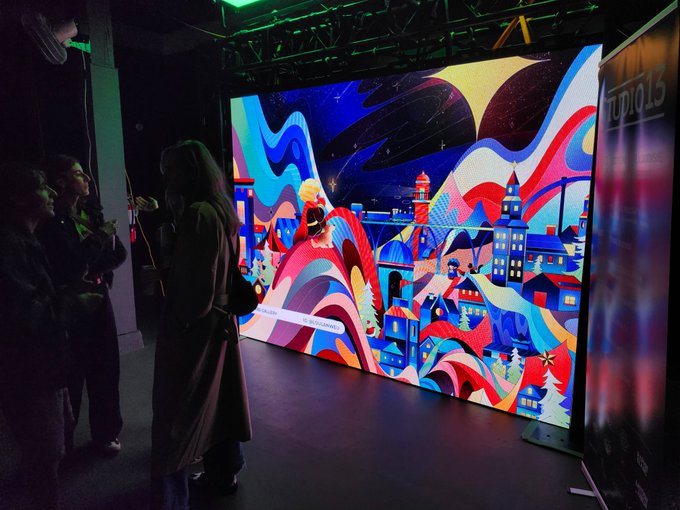 People stand in front of a large, vibrant interactive digital display featuring a colorful, abstract cityscape with the text 'STUDIO 13' visible on a nearby screen.