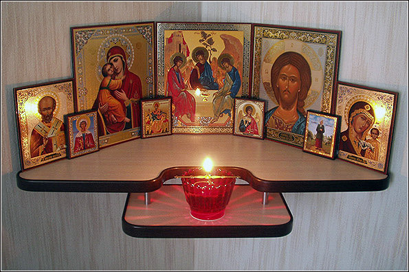 An Orthodox Christian icon corner featuring multiple religious icons including the Virgin Mary, Christ, and various saints arranged on a corner shelf with a red votive candle glowing in the center.