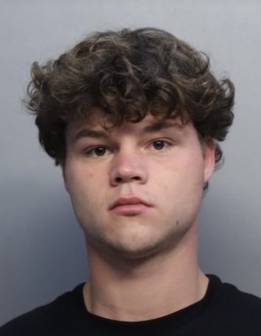 A mugshot-style portrait of a young man with curly brown hair wearing a black shirt against a plain gray background.