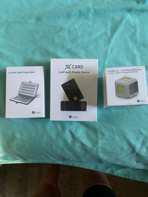 Three Ellipal hardware cryptocurrency wallet product boxes are displayed on a teal fabric surface, featuring the "ELLIPAL Seed Phrase Steel," "X CARD - Card Vault: Simple, Secure," and "ELLIPAL Joy - Seed Phrase Generator," presented as a celebratory gift set.