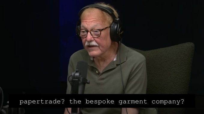 An elderly man with glasses and headphones sits in a podcast studio, speaking into a microphone while a caption reads "papertrade? the bespoke garment company?"—suggesting he is questioning or discussing an expensive tailor service referenced in the accompanying meme about men overspending on clothing instead of therapy.