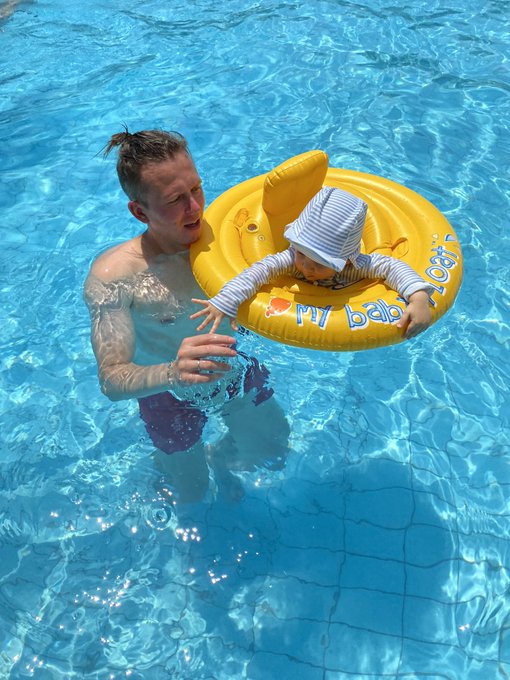 A man in a swimming pool supports a baby wearing a striped sun hat in a yellow "My Baby Float" swim ring.