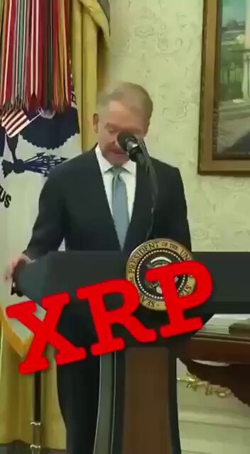 The image features a man standing at a podium with his hands on his hips, giving a speech or presentation. The text "XRP" can be seen in the image, which suggests that there might be an event related to cryptocurrency taking place. The man is wearing a suit and tie, indicating a formal setting for the occasion. Additionally, there are multiple flags visible in the background, further emphasizing the importance of this gathering or event.