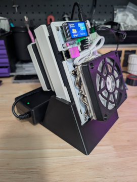 A DIY electronic device features a small LCD screen, a cooling fan, and circuit boards mounted in a black stand on a wooden desk.