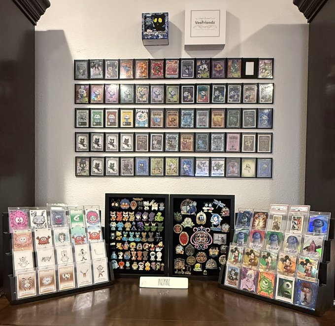A wall display of trading cards featuring the VeeFriends brand, with rows of holographic cards mounted on the wall and pin collections displayed on stands below.