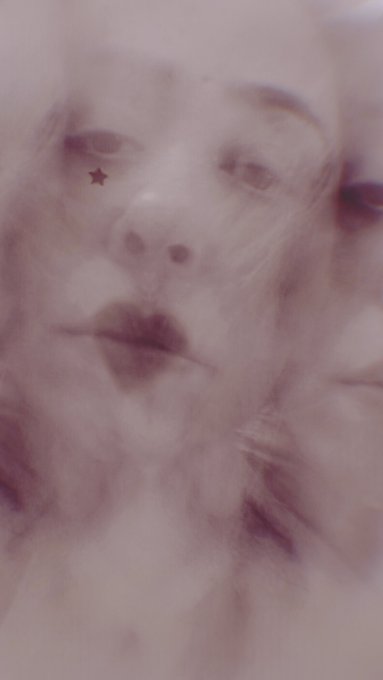 A dreamy, blurred close-up portrait of a person's face with a small star drawn near the eye, rendered in soft pink and purple tones.