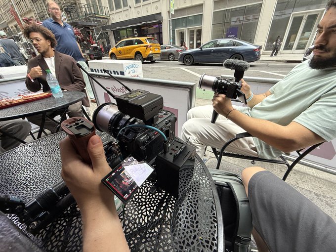 The image shows a group of people sitting at an outdoor cafe, with one person holding a camera while another has a microphone. The scene appears to be set in a city environment where there are cars parked nearby. A caption below the photo reads "THE ETHEREUM FOUNDATION CARES ABOUT BUILDERS". This suggests that the people present might be involved in some form of media project or event related to urban development, architecture, or community issues.
