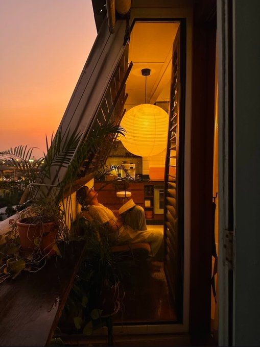 A moody, warm-toned photograph of a person reading a book in a cozy room with a large glowing paper lantern, viewed through an open window or door against a pink sunset sky.