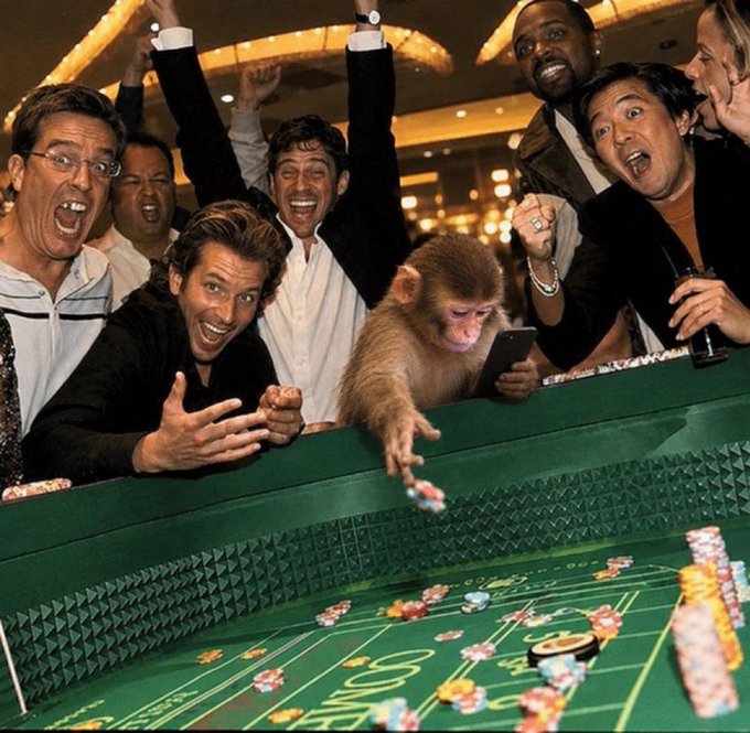 A group of excited men cheer at a craps table while a monkey reaches out to toss casino chips onto the green felt.