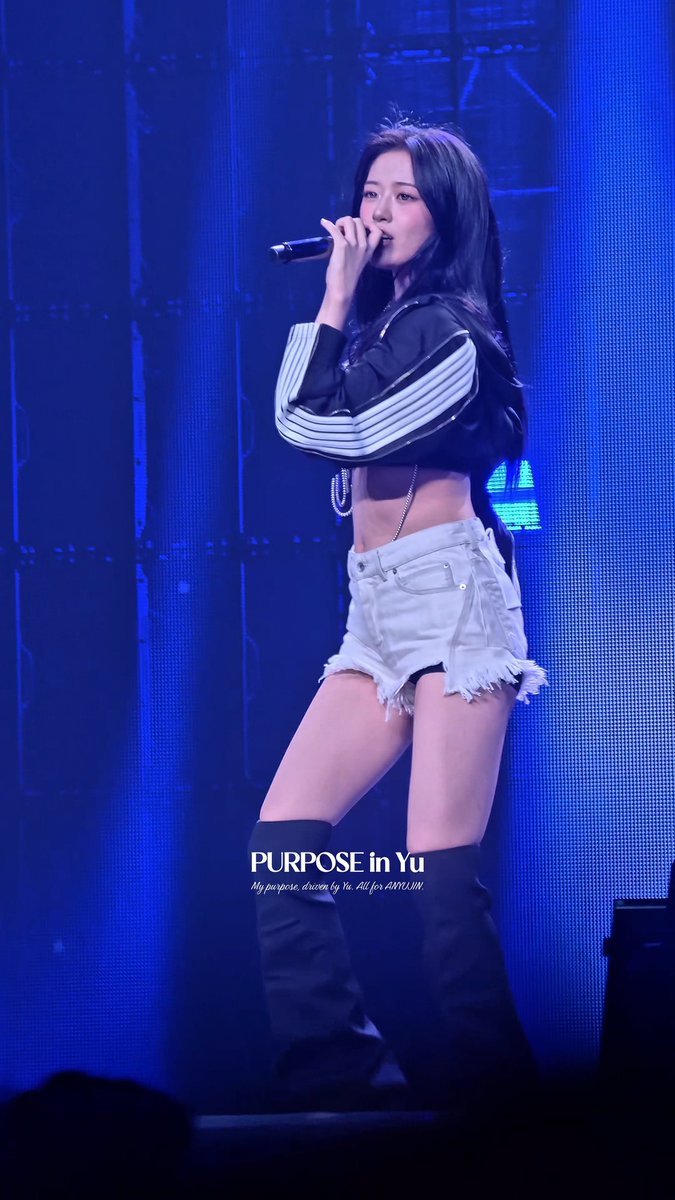 A concert photo of a female performer singing into a microphone, wearing a black and white striped cropped jacket and white denim shorts, with text overlay 'PURPOSE in Yu - My purpose, driven by Yu. All for ARIRANG.'