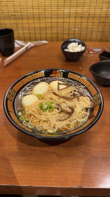 A bowl of ramen noodles with boiled eggs and green onions in a decorative bowl, served on a wooden table alongside smaller side dishes.