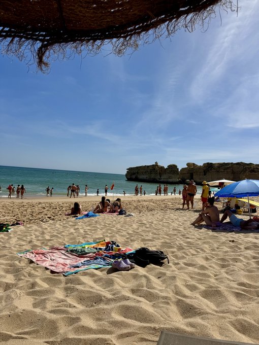 A sunny beach scene with people relaxing on the sand, others wading in the turquoise water, and rocky cliffs in the distance under a blue sky.