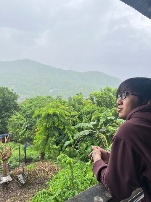 A woman with glasses gazes out over a misty, lush mountain landscape from a balcony, enjoying a peaceful rain-soaked view during a getaway from city noise. The serene, verdant scenery reflects the post's theme of a refreshing escape made possible by recent earnings, symbolizing the calm and reward of stepping away from busy life.