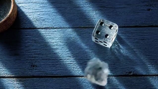 The image features a silver dice with black dots, which appears to be rolling across a wooden surface. There are also two small rocks visible near the bottom of the frame. The background suggests that this scene takes place outdoors or in an open area where sunlight is shining on the objects.