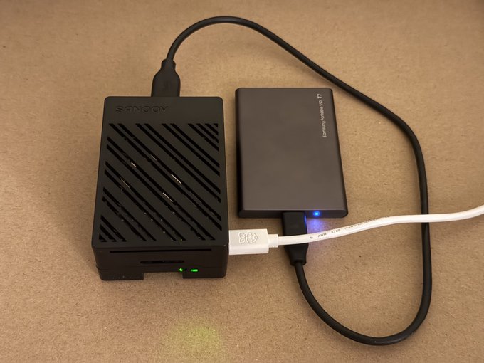 A SanDisk Rage R drive and a Raspberry Pi 5 sit connected on a wooden surface, with the drive's blue LED lit indicating active operation—showcasing the new Raspberry Pi 5 hardware being used to run the Umbrel Bitcoin Node IBD sync mentioned in the post.